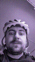 a selfie of a white person, wearing either headphones or a helmet, making an odd expression. the photo is edited to be colorized into a monochrome purple-ish color palette.